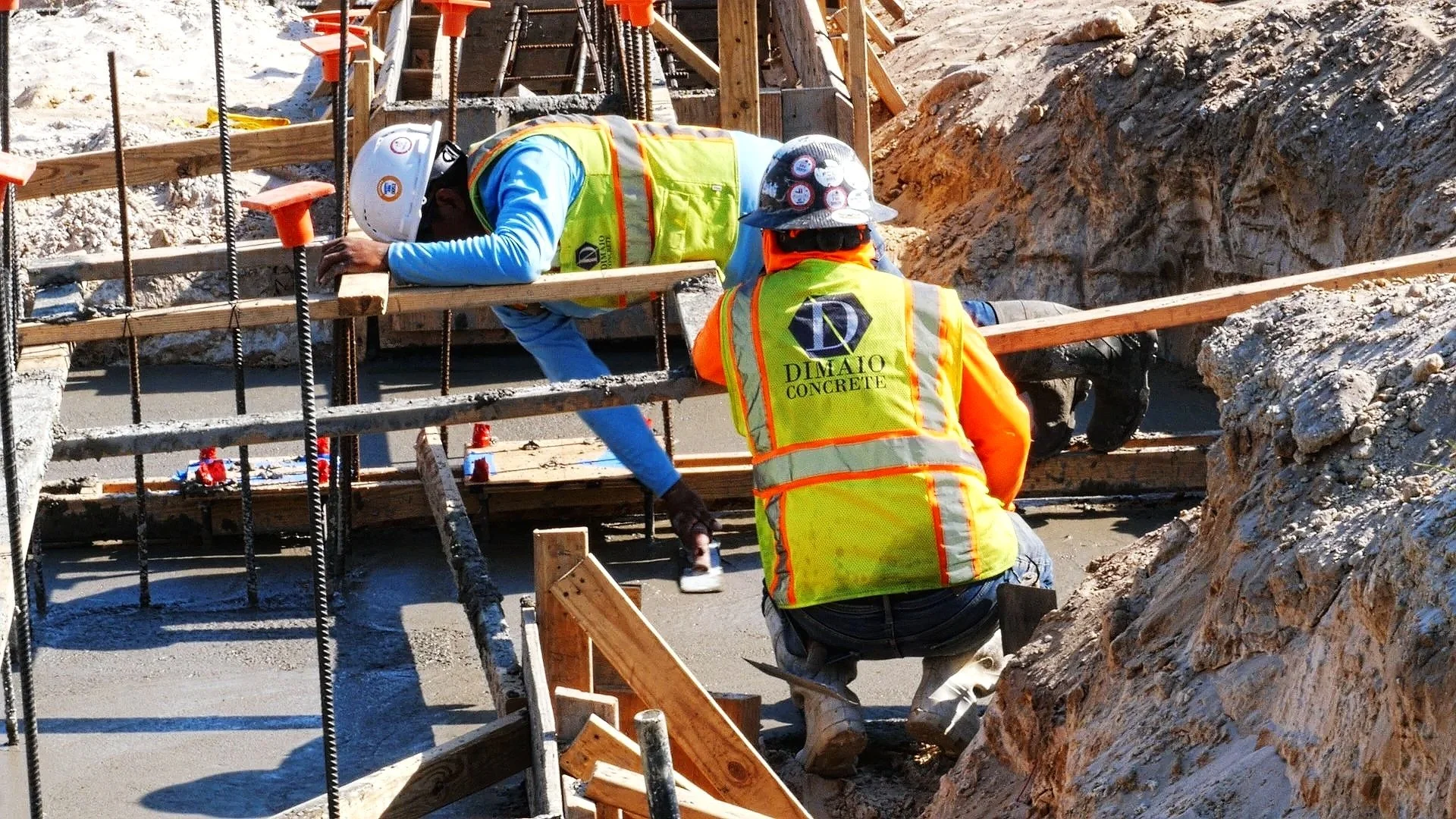 Construction workers wearing safety vests and helmets working on a concrete and rebar foundation at a construction site.