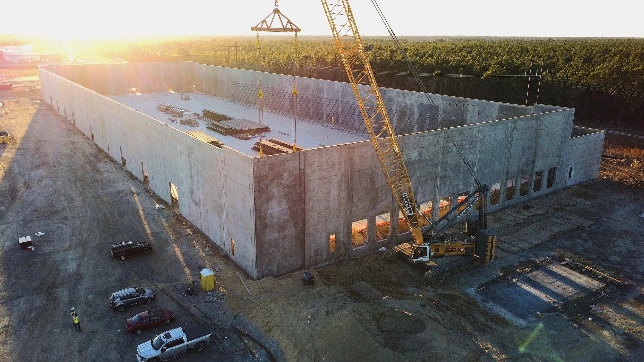 Construction site during sunset with a large concrete structure, a crane lifting materials, and several parked vehicles.