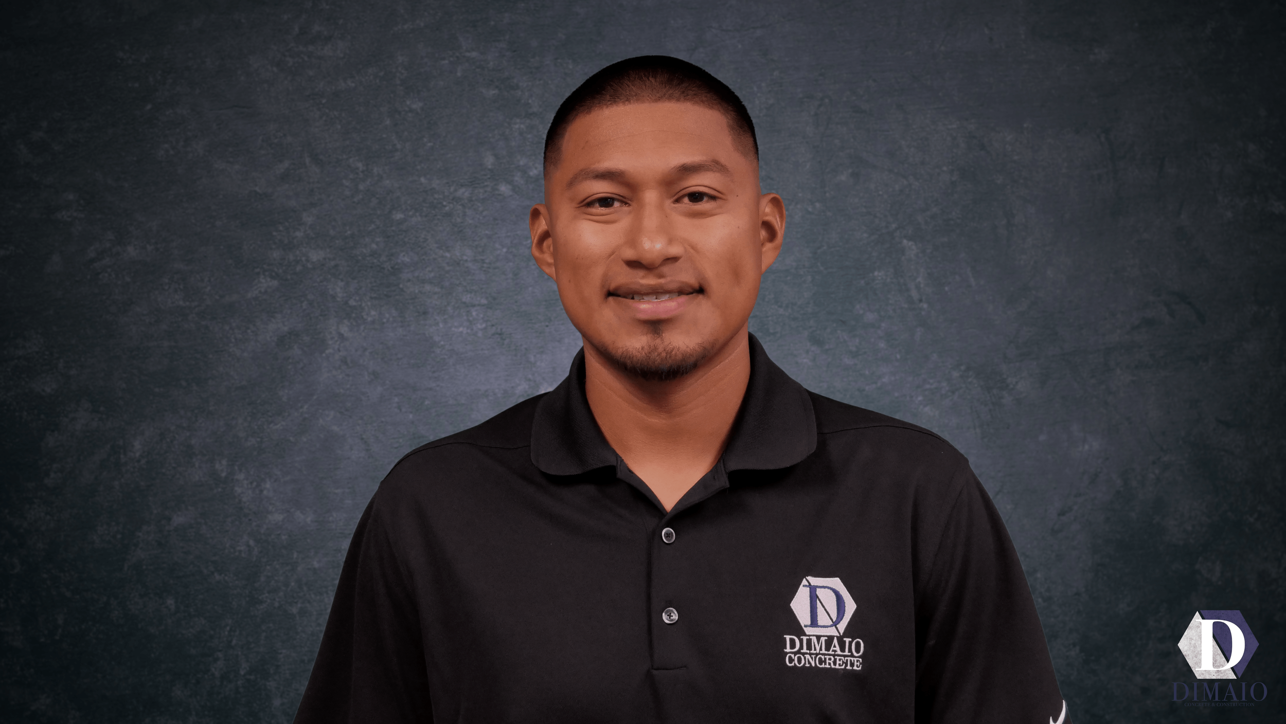 Portrait of a young man with short hair, wearing a black collared shirt with a logo on the chest that reads 'DIMAIO CONCRETE' against a dark textured background.