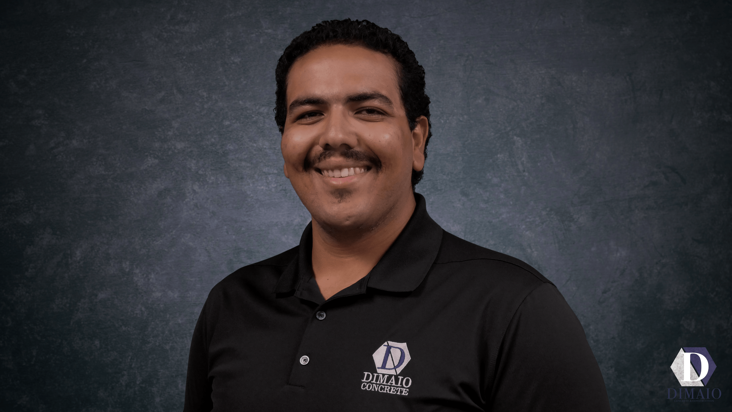 Portrait of a man with dark curly hair, mustache, and goatee, wearing a black polo shirt with a logo that says 'Dimao Concrete' on the chest, standing in front of a dark textured background.