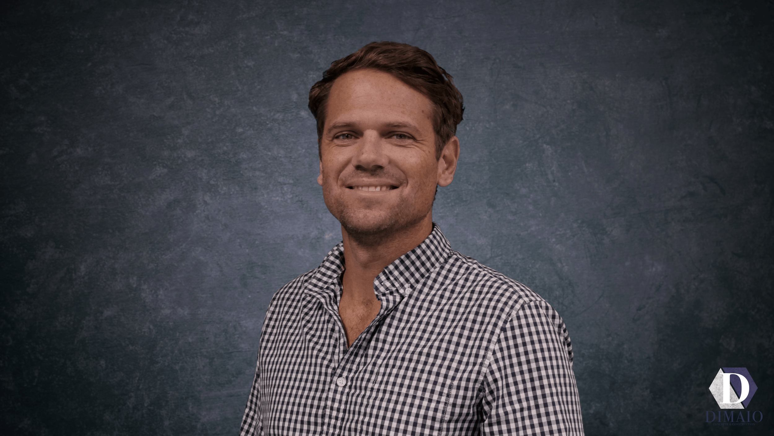 A young man with brown hair wearing a black and white checkered shirt, smiling slightly, standing in front of a dark textured background.