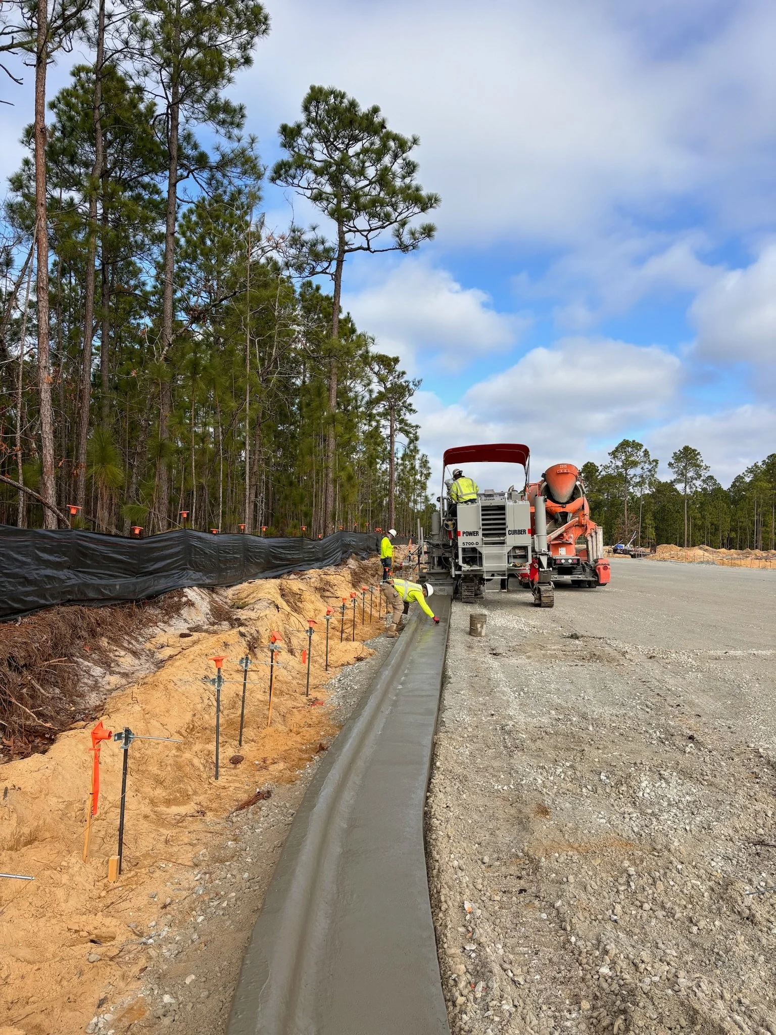 Concrete curb being constructed by a slipform paving machine on a construction site with red safety fencing and construction worker nearby.