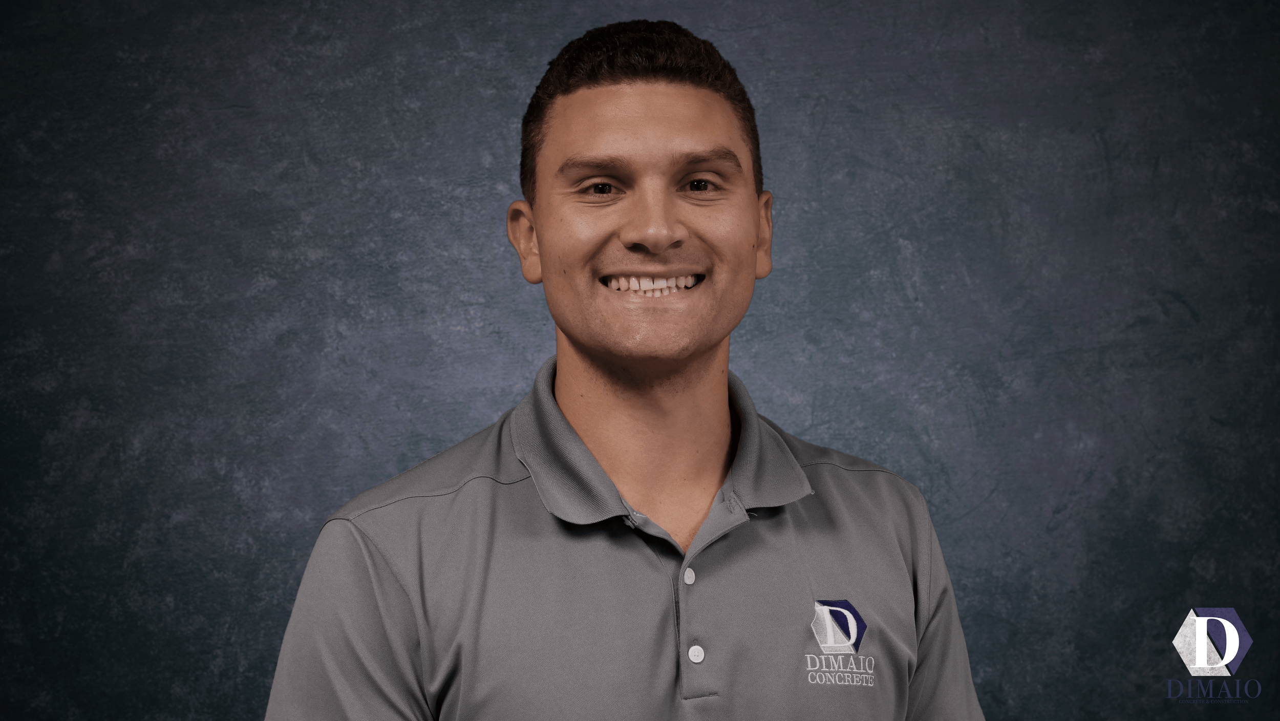 A young man with short brown hair smiling at the camera, wearing a gray collared shirt with a logo that reads "Dimaio Concrete," standing against a dark textured background.