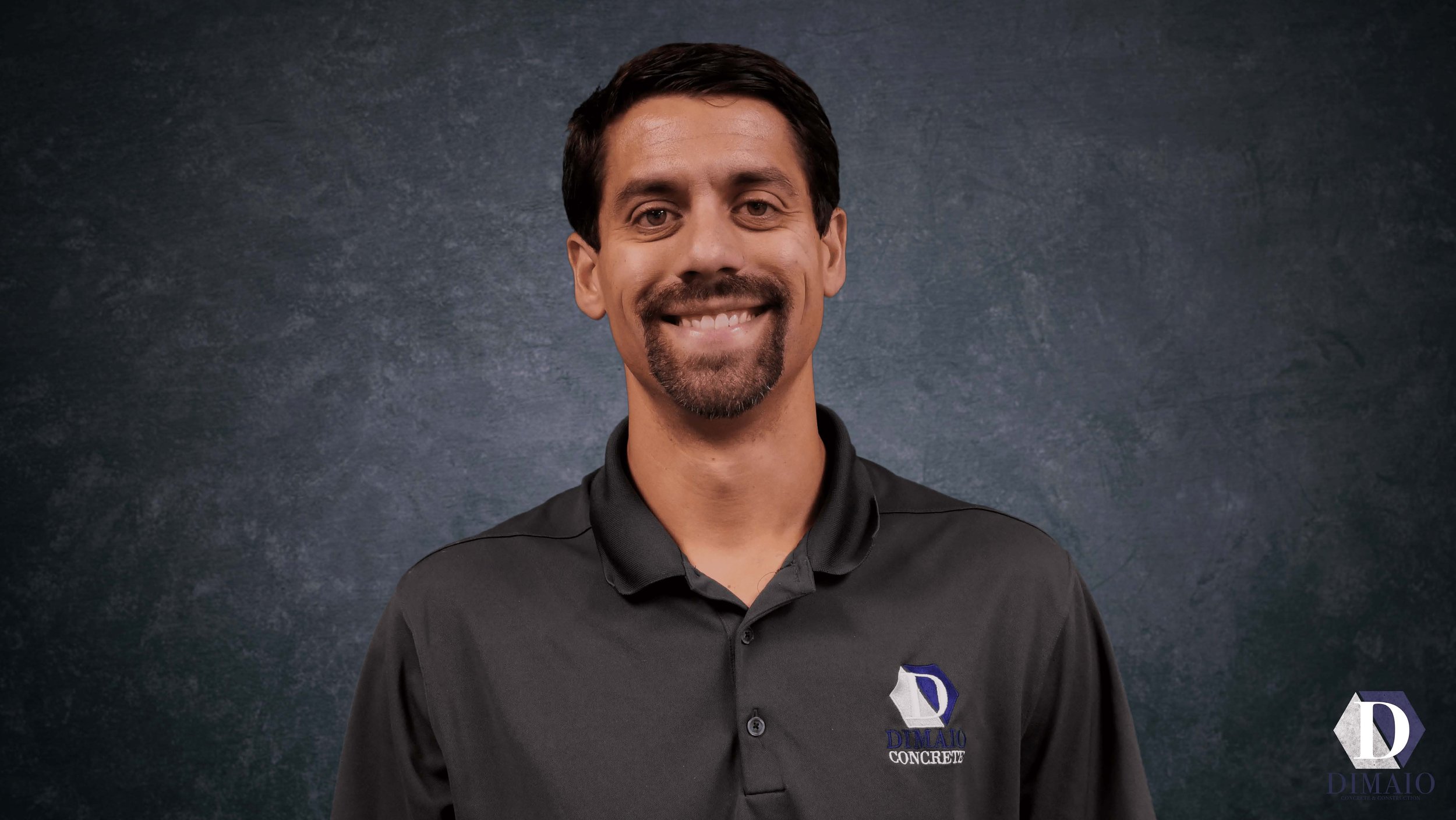 A man with dark hair and a goatee smiling, wearing a black collared shirt with a logo that reads 'DIMAO CONCRETE' and a letter 'D' emblem, standing against a dark, textured background.