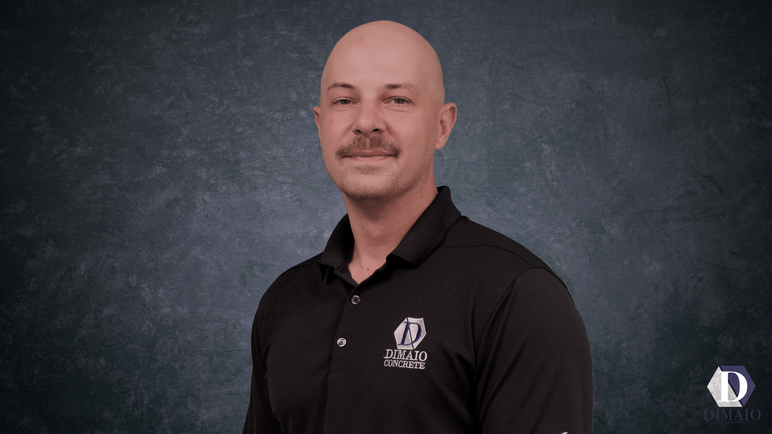 Headshot of a bald man with a mustache and goatee, wearing a black collared shirt with a logo that reads "DIMAIO CONCRETE" on the left chest, standing against a dark textured background.