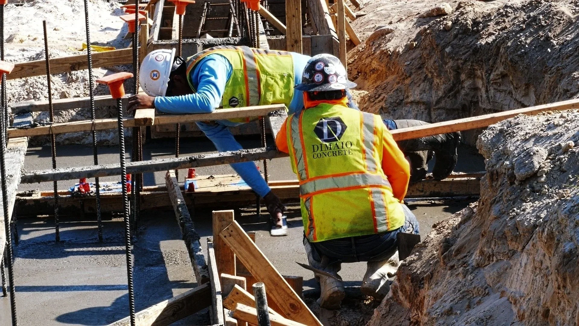 Two construction workers in safety vests working on a concrete foundation with rebar at a construction site.
