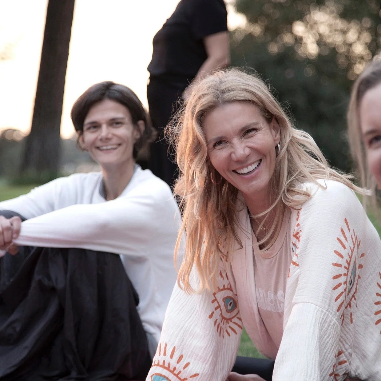 Group of smiling women outdoors during sunset, sitting on grass with trees in background, enjoying each other's company.