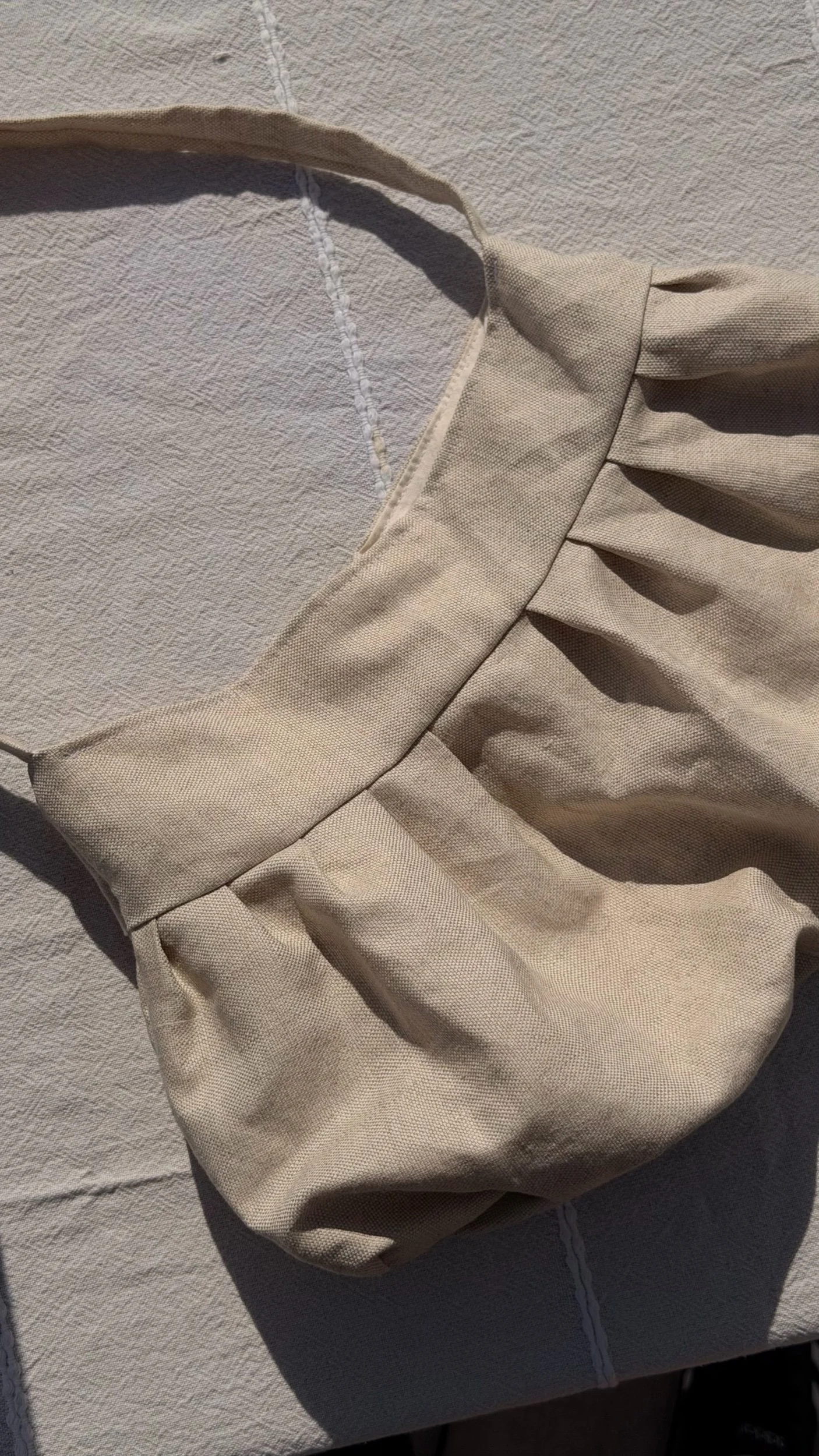 A beige fabric purse with pleats and an open top, placed on a textured light-colored surface.