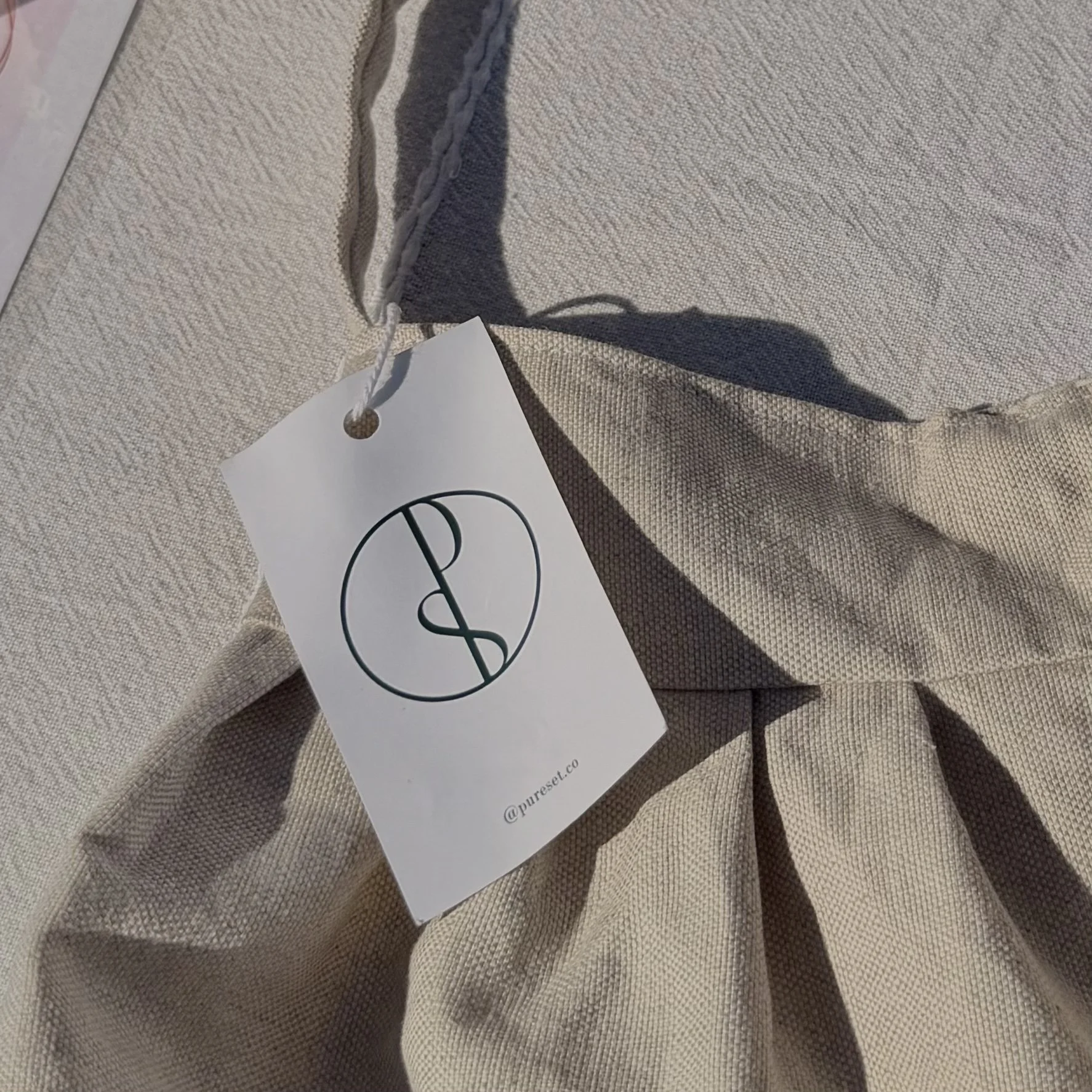 Close-up of a beige fabric garment with a white tag attached, featuring a black circular logo with intertwined initials and the text '@pureset.co'.