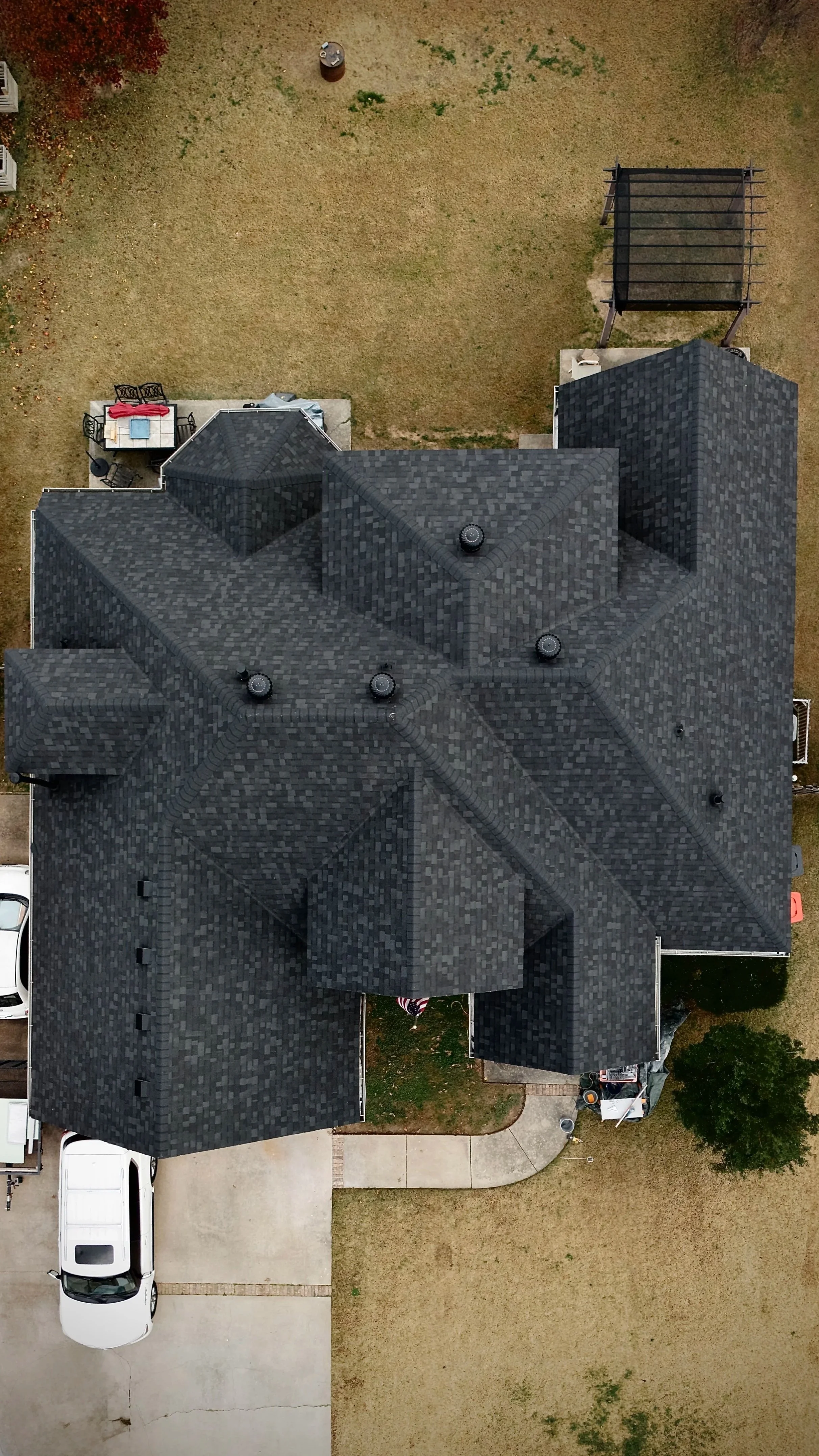 Aerial view of a large black shingled house with a front porch, parking space with a white van, outdoor patio furniture, a small grassy yard, and some trees.
