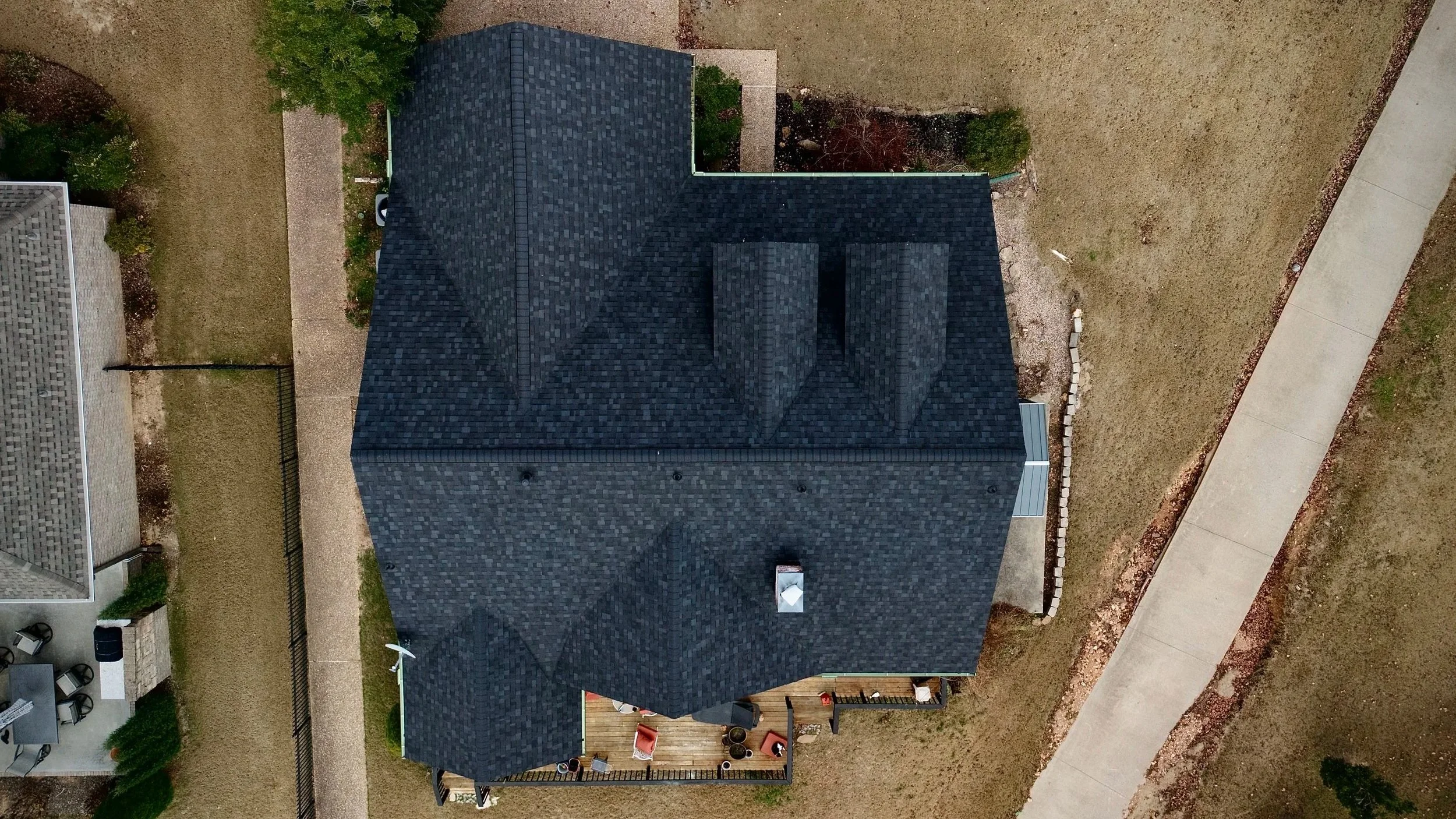 Aerial view of a black shingled house with a backyard deck area. The deck has outdoor furniture, including chairs and a table, and is surrounded by a grassy yard with a curved walkway. The neighboring houses and yards are partially visible.