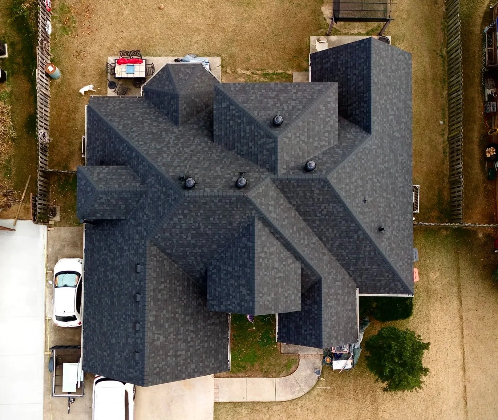 Aerial view of a large house with a dark black shingle roof, surrounded by neighboring houses, small yards, and driveways. The backyard has a patio with outdoor furniture, a small grassy area, and a few trees.