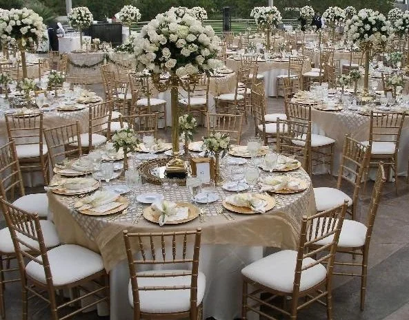 Elegant outdoor event setup with round tables decorated with white tablecloths, gold Chiavari chairs, and large floral centerpieces of white flowers.