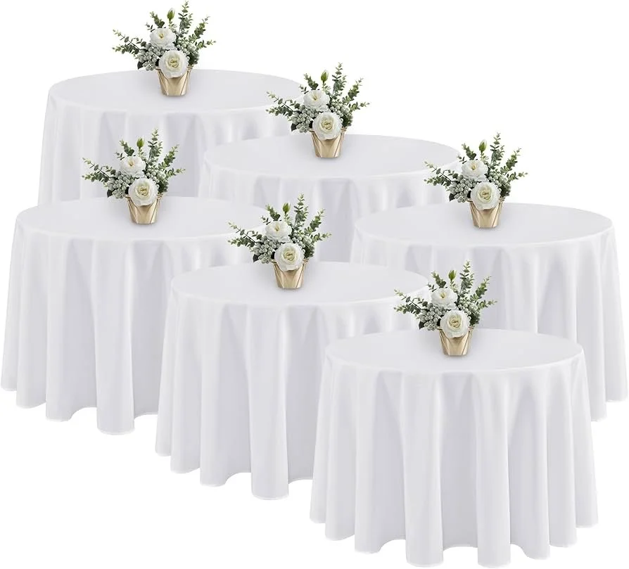 Six round tables covered with white tablecloths, each decorated with a small floral arrangement in a gold vase, featuring white flowers and greenery.