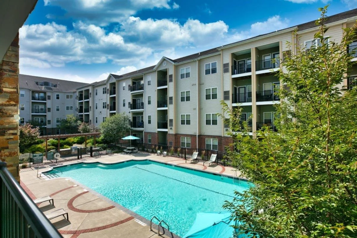 Outdoor swimming pool in the courtyard of a multi-story apartment complex with trees and a blue sky.