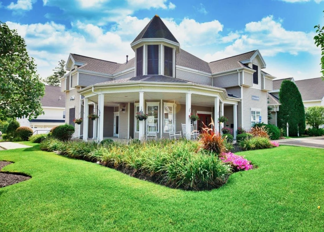 Large Victorian-style house with a turret, wraparound porch, boxing chairs, and hanging flower baskets, surrounded by a well-manicured lawn and garden with colorful flowers.