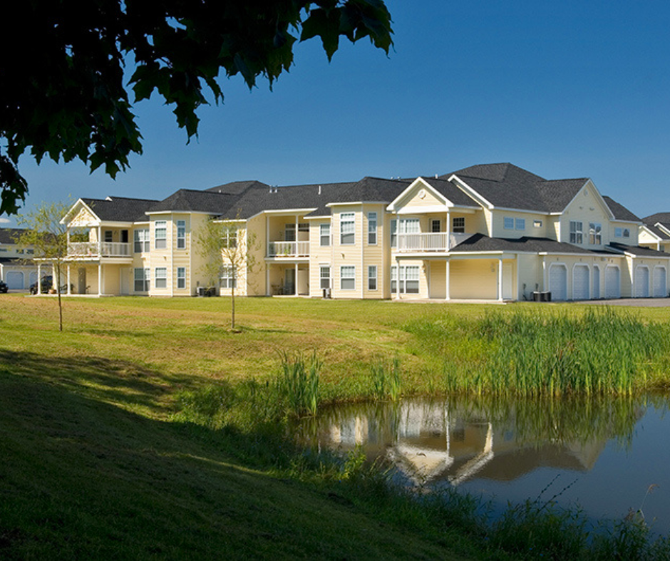 A large, yellow multi-unit residential building with balconies and garages situated on a grassy lawn next to a pond, under a clear blue sky.