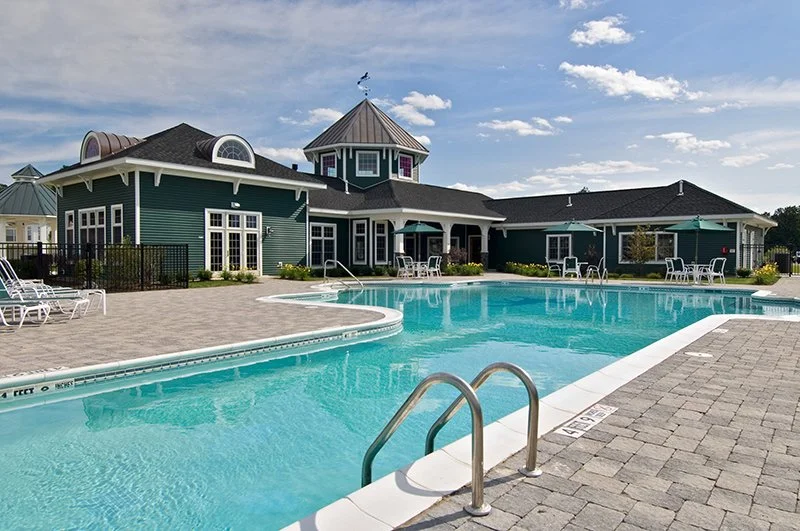 Outdoor swimming pool with lounge chairs and a large house with multiple roof styles and green exterior in the background under a partly cloudy sky.