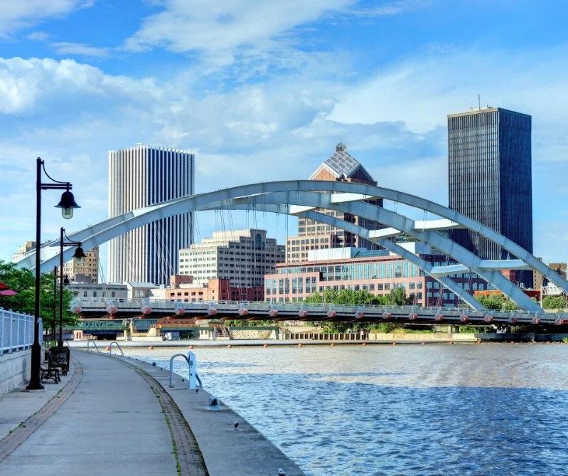 City skyline with modern glass buildings, a white arched bridge over a river, and a walkway with street lamps in the foreground.