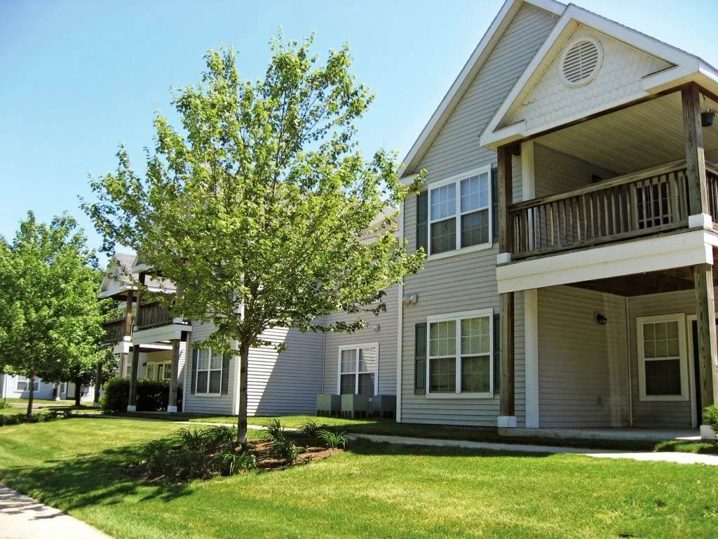 Multi-story residential buildings with balconies, green grass, and trees in a suburban neighborhood.