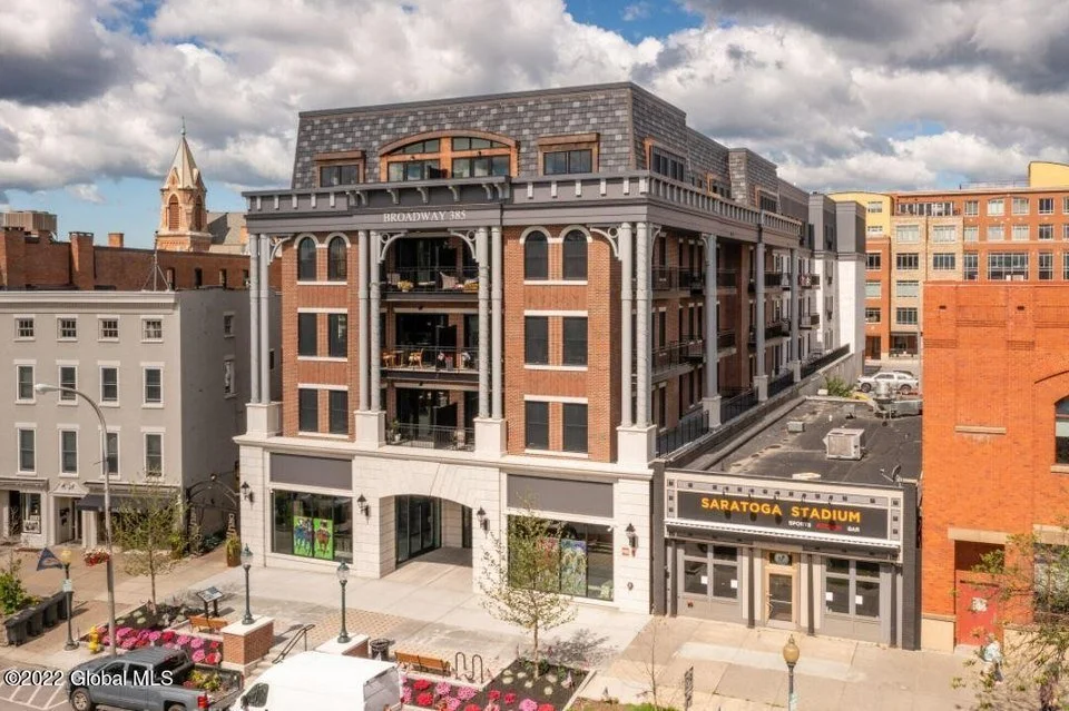 Multi-story brick building with balconies, labeled Broadway 385, adjacent to Saratoga Stadium sports arena, on a city street with pedestrians, trees, and parked cars under a partly cloudy sky.