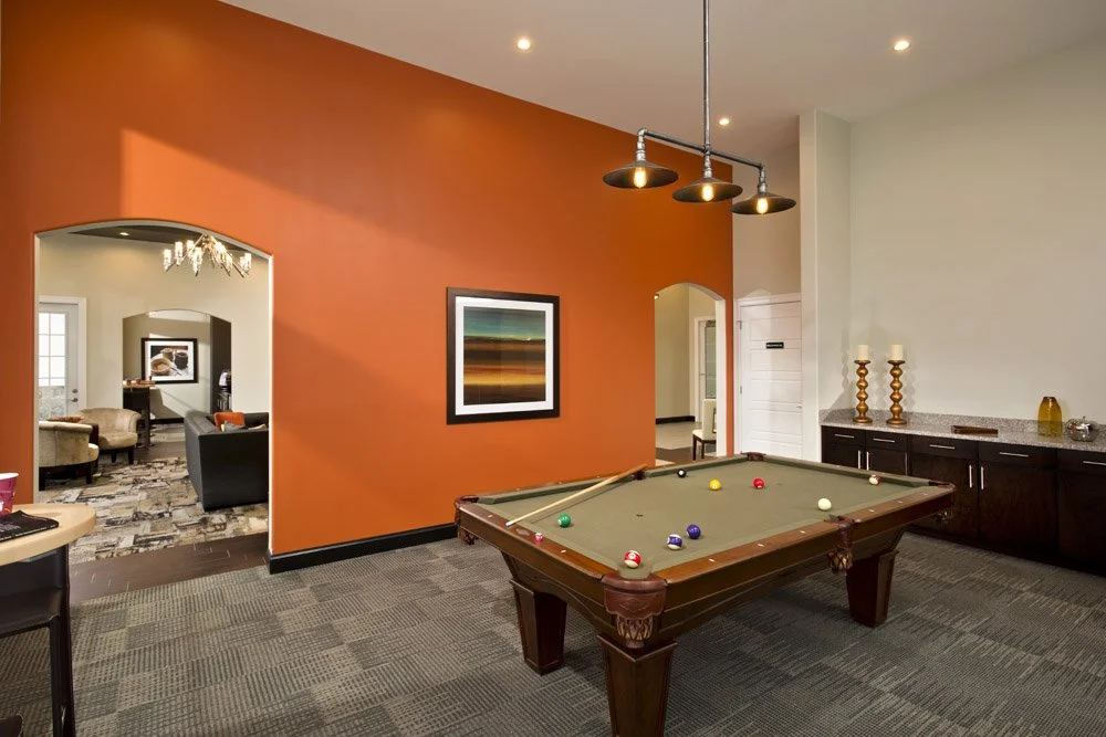 An indoor room with a pool table, orange accent wall, and a sideboard with candles. There is an opening to a sitting room with chairs and a chandelier visible.