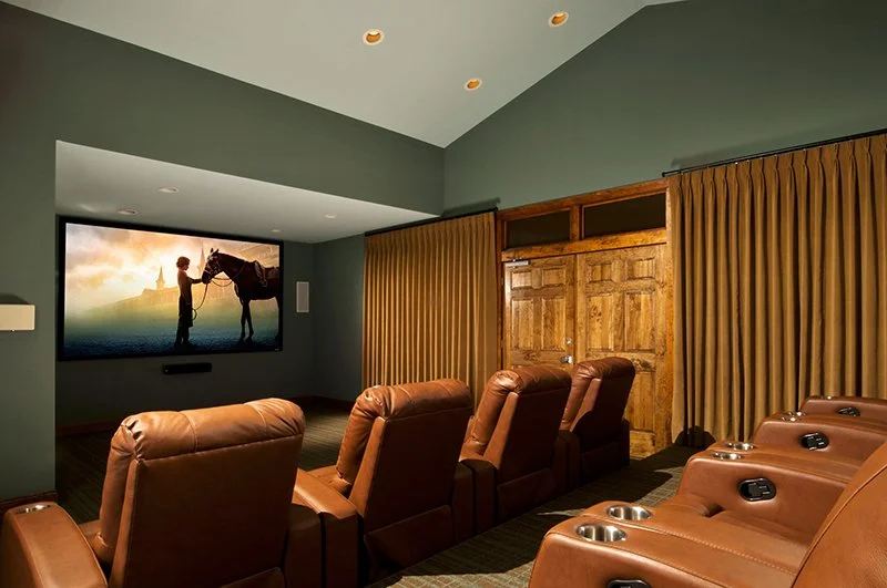 A home theater room with leather recliner seats facing a large flat-screen television showing a scene from a movie with a child and a horse. The room has green walls, a wooden door, and gold curtains.