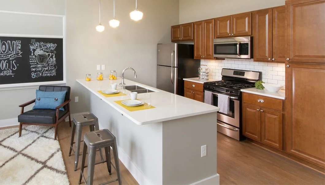 Modern kitchen with wooden cabinets, stainless steel appliances including refrigerator, microwave, and stove, white subway tile backsplash, granite countertops, small bar area with yellow placemats and white dishes, black chair with blue pillow, chal