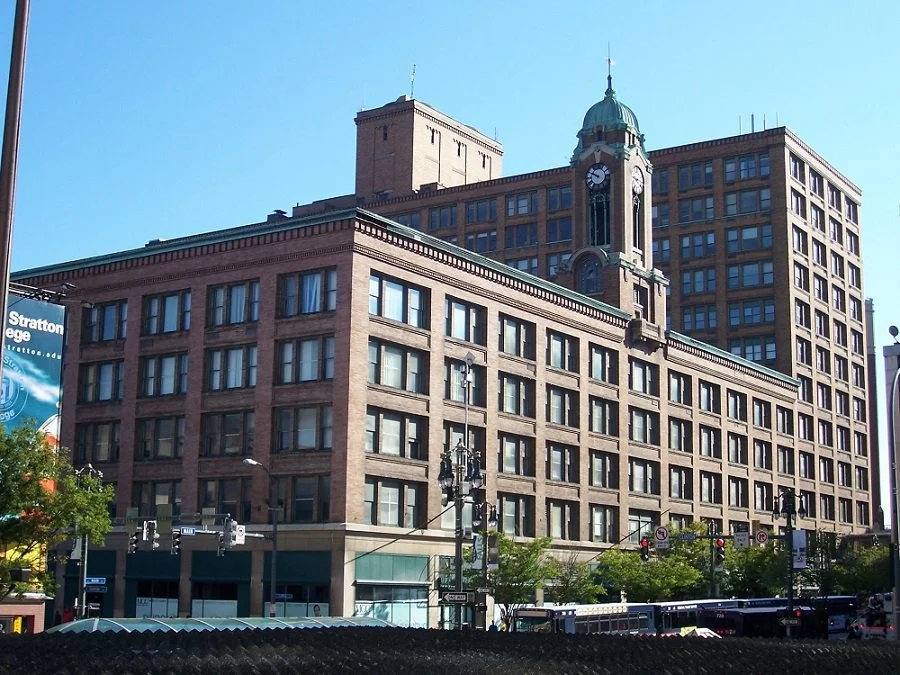 A large historic brick building with a clock tower in an urban area, with surrounding trees, traffic lights, and streetcars visible in the foreground.