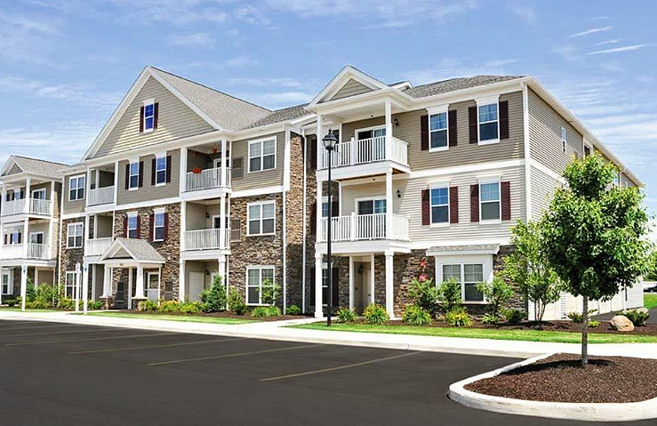 Exterior view of a multi-story residential apartment building with balconies, surrounded by a parking lot and landscaped greenery, under a blue sky.
