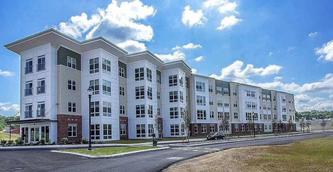Large modern apartment building with multiple floors, white exterior, numerous windows, and a brick base, under a partly cloudy blue sky.