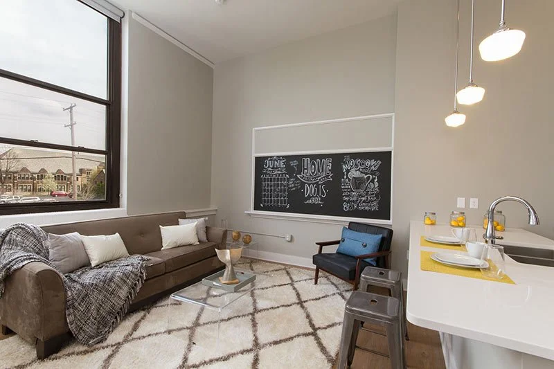 Modern living room with large window, beige sofa with pillows and blanket, black chalkboard wall with writing, blue armchair, and a kitchen island with yellow accents.