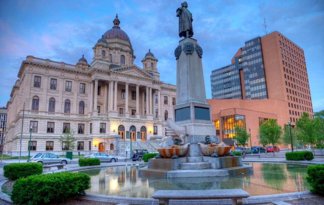 City square with a historic courthouse, a statue with water features, and modern office buildings under a blue sky.