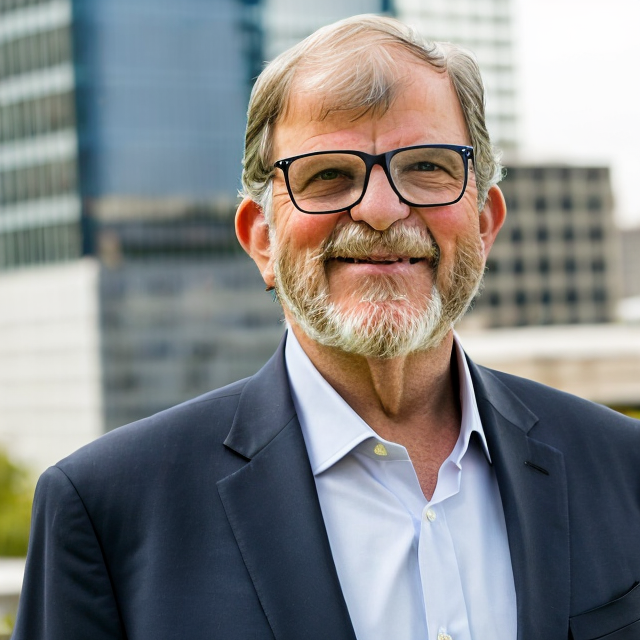Portrait of a smiling middle-aged man with glasses and a beard, wearing a suit jacket and a white shirt, standing outdoors in an urban setting with modern office buildings in the background.