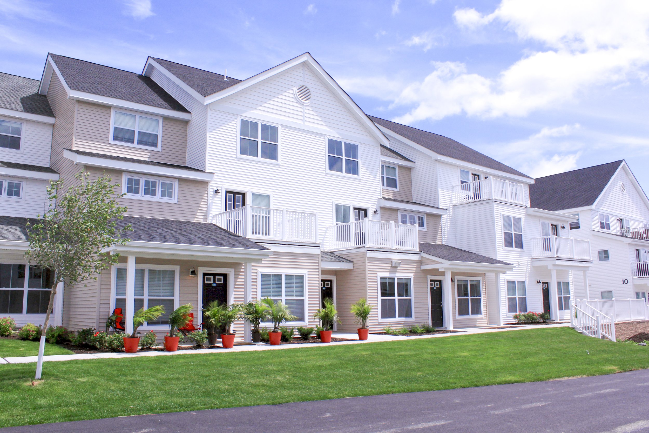 Modern white multi-story apartment complex with balconies, potted plants, and a green lawn under a blue sky.