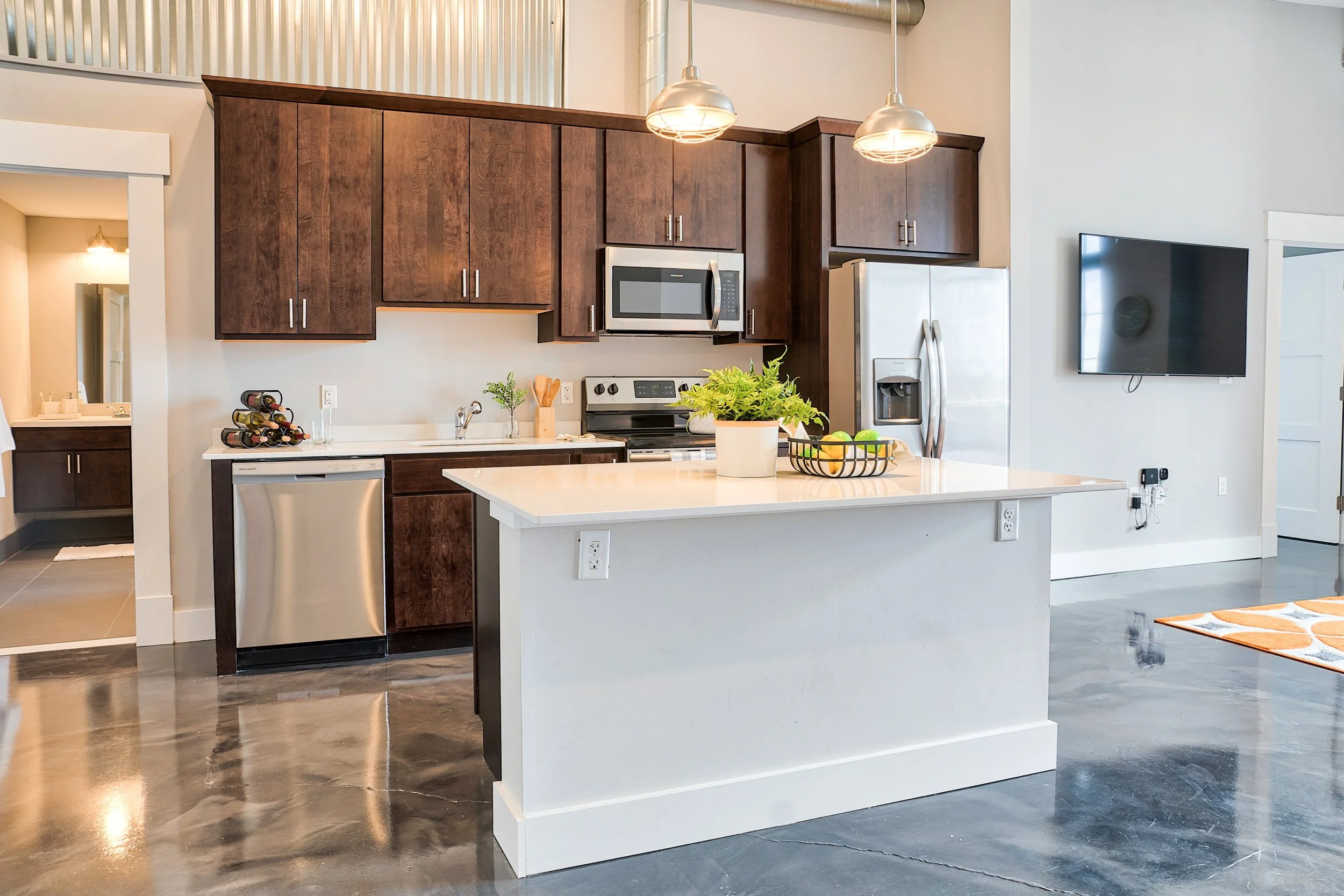 Modern kitchen with white island, dark wooden cabinets, stainless steel appliances, potted plant, and wall-mounted television.