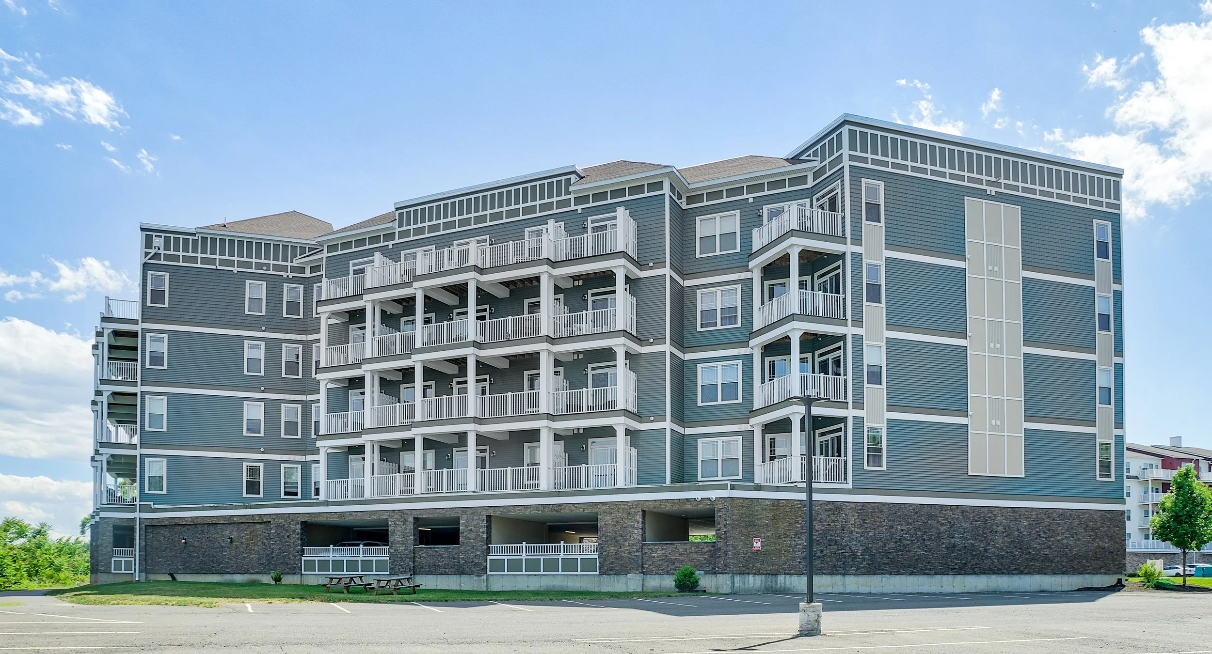 Multi-story residential building with blue siding and white balconies, parking lot in front, and a small tree on the right