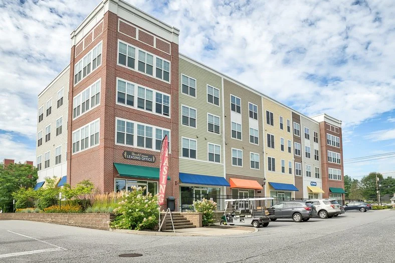Multi-story mixed-use building with retail storefronts on the ground floor and residential units above, with parked cars in front and a partly cloudy sky.