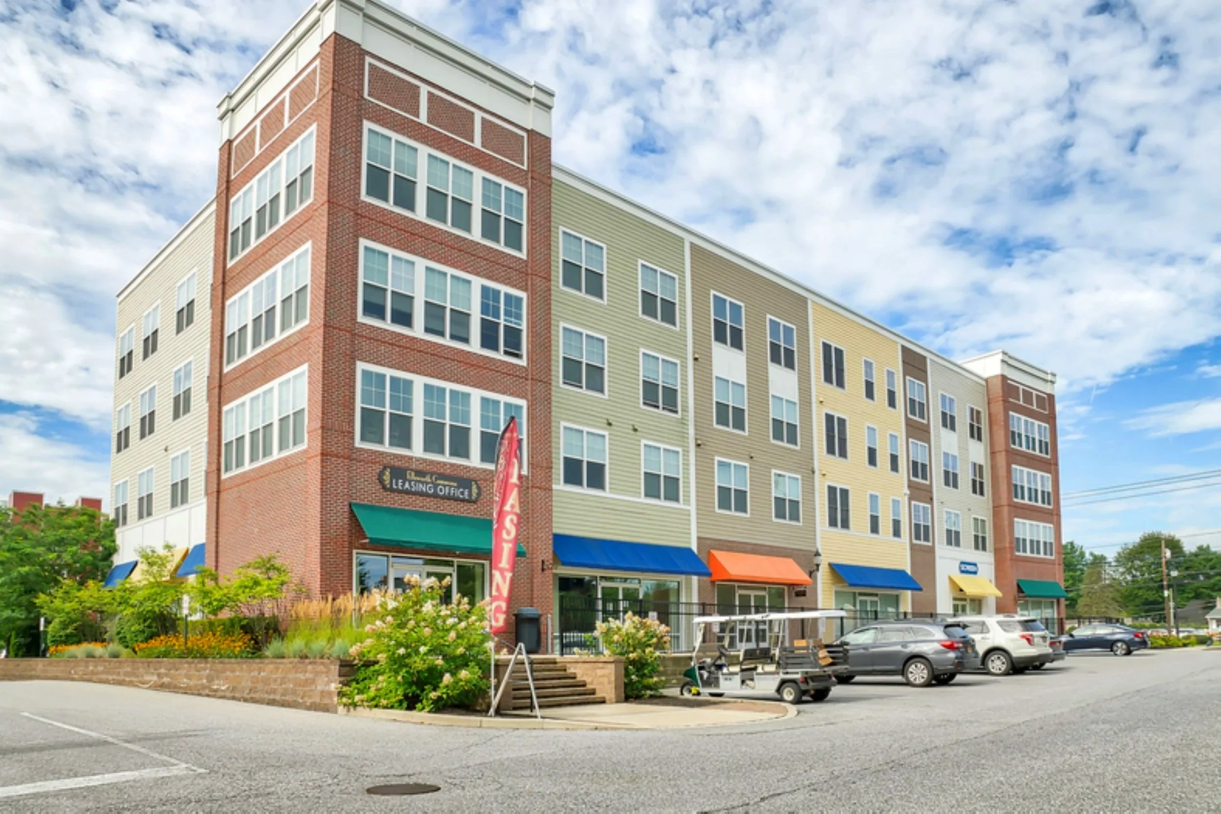 Multi-story commercial building with retail storefronts, parked cars, and a leasing office sign, under a cloudy blue sky.