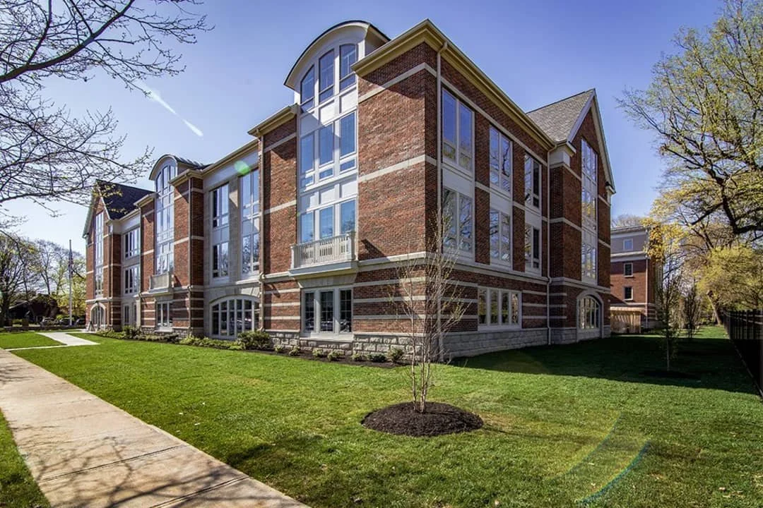 A multi-story brick apartment building with large windows and a small front lawn with a sidewalk, trees, and a clear blue sky.