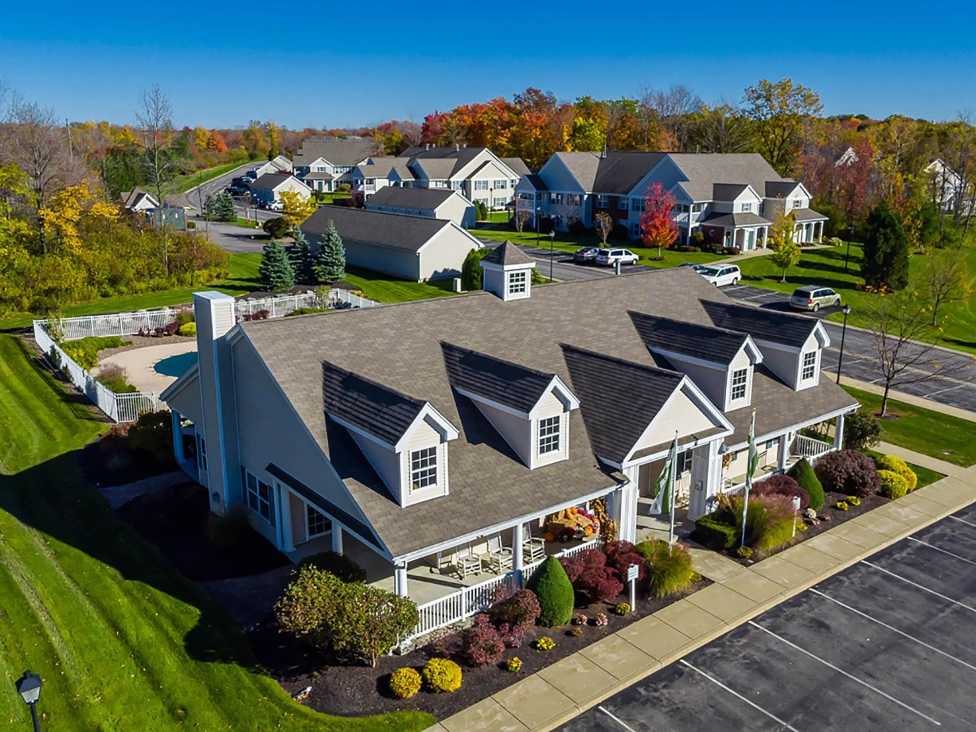 Aerial view of a suburban neighborhood with a large white building, parking lot, playground, and surrounding houses with fall foliage.