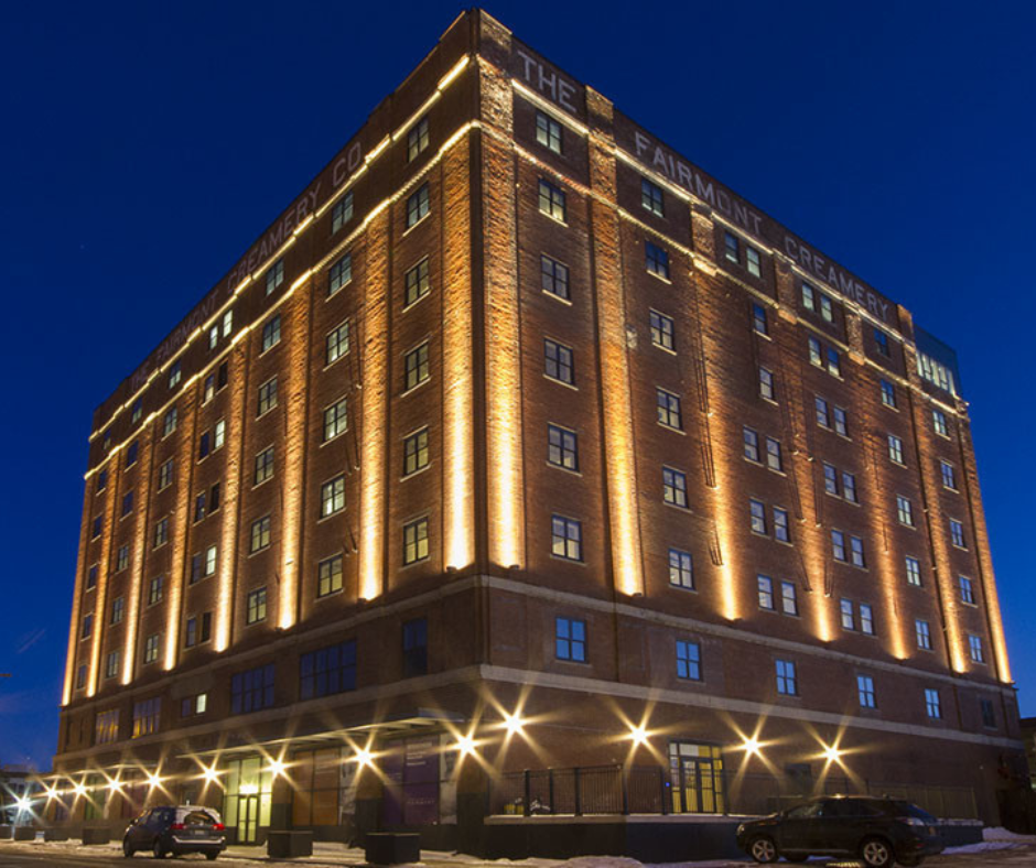 Night view of a tall brick building with illuminated vertical lines and signs reading "The Fairmont Creamery Company" at the top. Car parked in front.