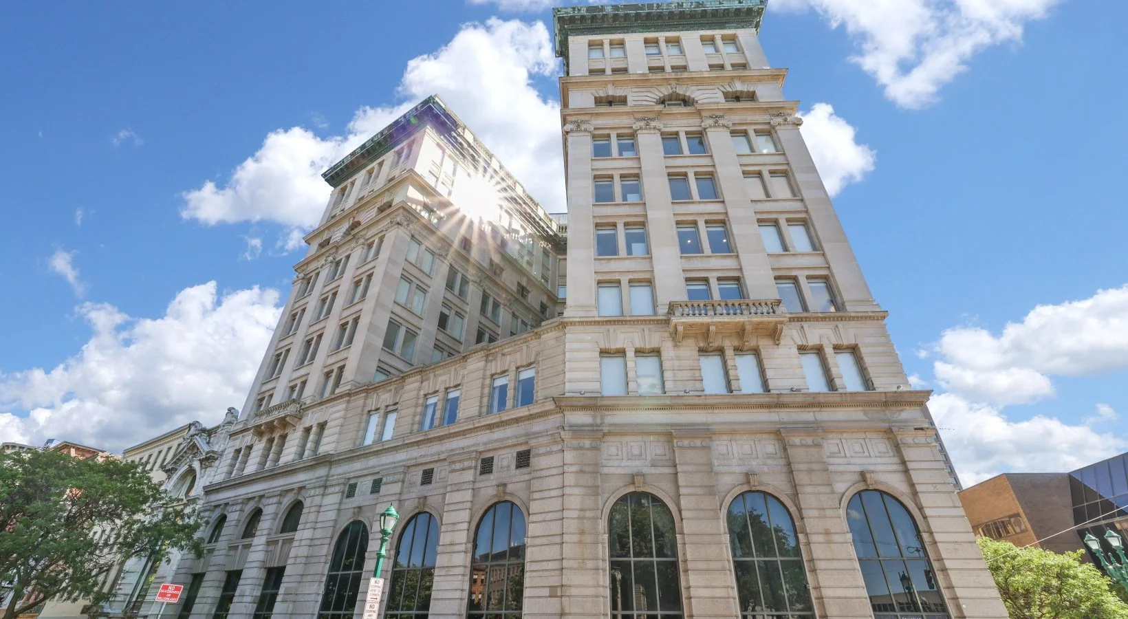 A tall historic building with arched windows at the bottom and rectangular windows above, set against a partly cloudy blue sky with sunlight shining, and a few trees at the street level.