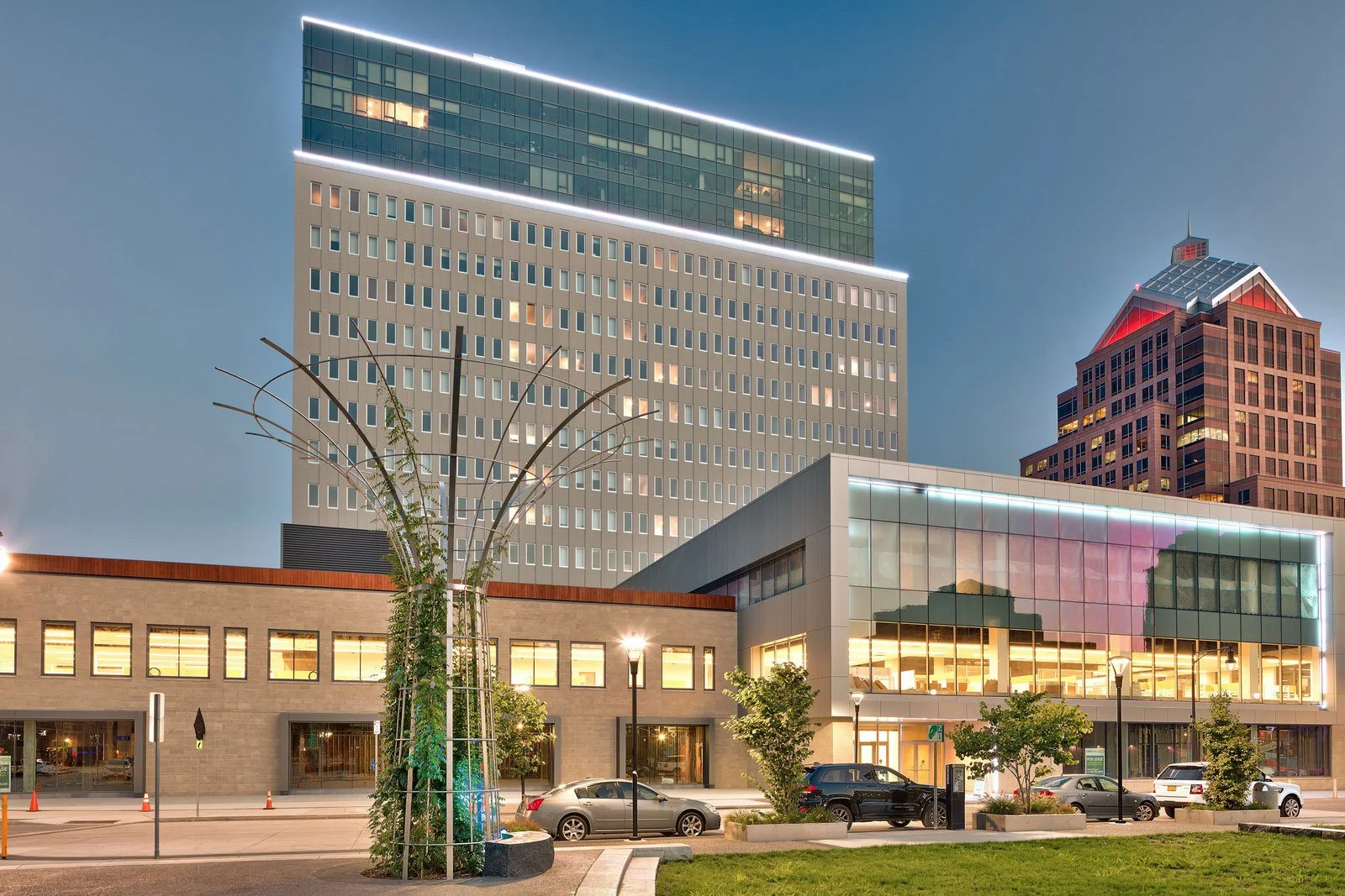 Urban scene at dusk featuring modern multi-story office buildings with glass and concrete facades, parked cars, streetlights, trees, and a landscaped sidewalk.