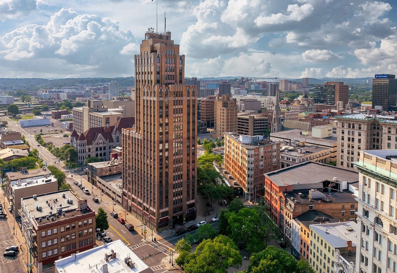 Aerial view of downtown cityscape with tall buildings, green trees, and streets under a partly cloudy sky.