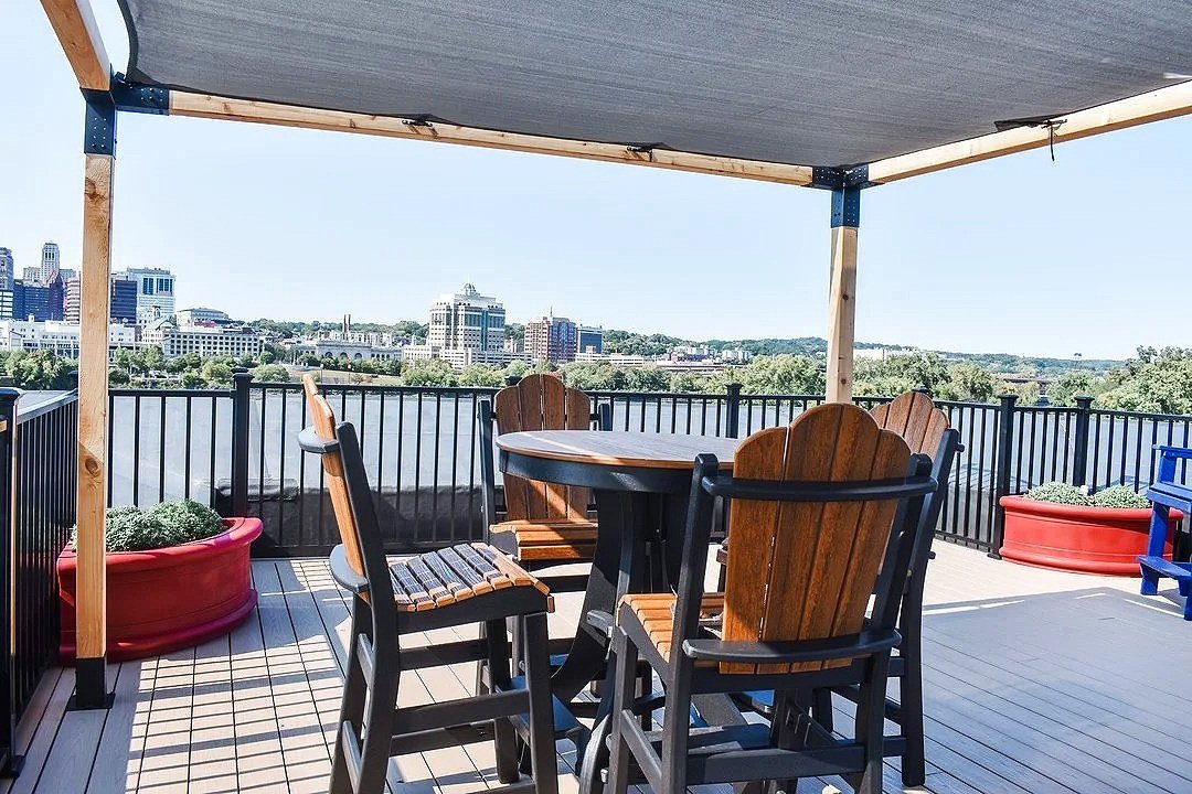 Outdoor rooftop patio with a round table, wooden and black chairs, potted plants, and a city skyline view under a canopy.