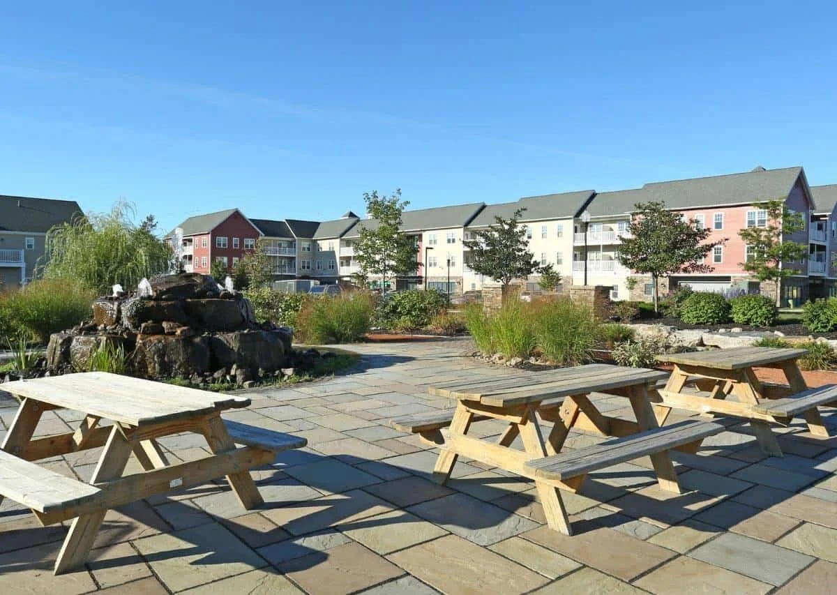 A park with wooden picnic tables, a rock fountain with water on top, and green plants, with multi-story apartments in the background under a clear blue sky.