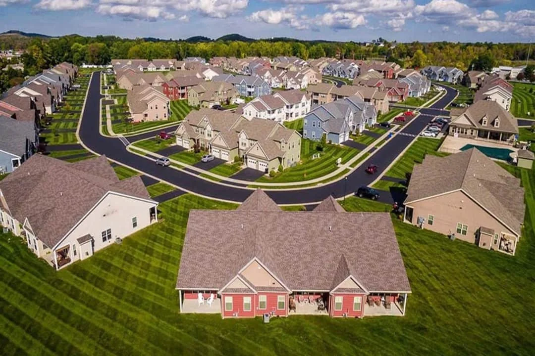 Aerial view of a suburban neighborhood with rows of single-family houses, green lawns, paved roads, and some parked cars, under a partly cloudy sky.