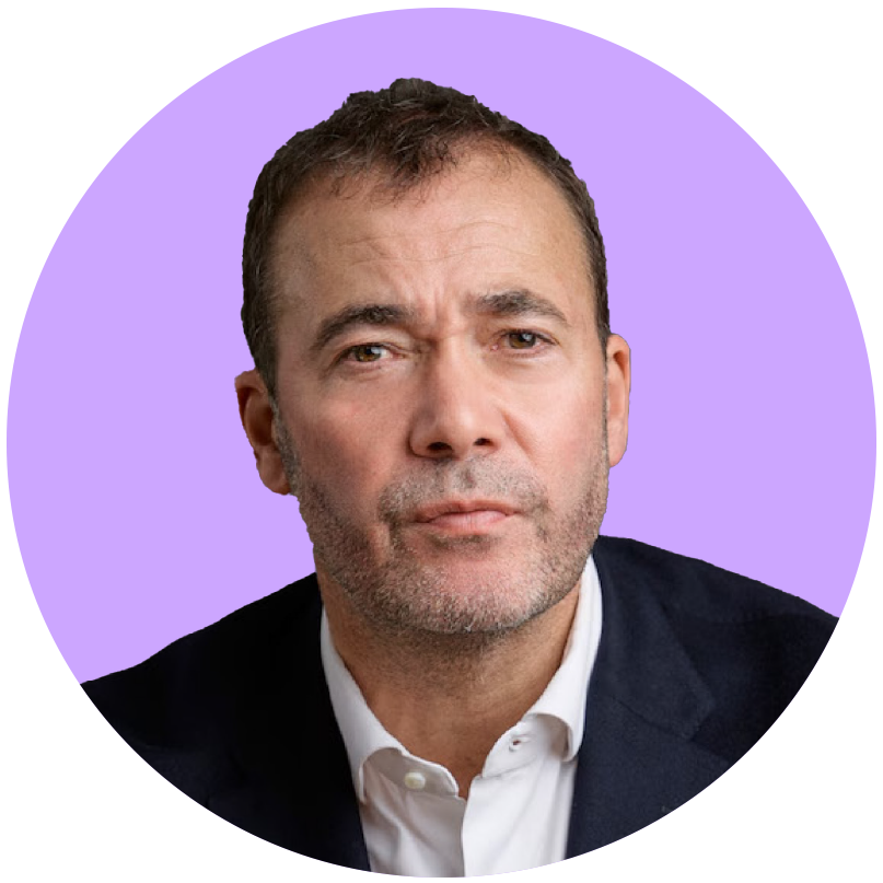 Close-up image of a middle-aged man with short brown hair, gray stubble, wearing a black blazer and white shirt against a purple background.