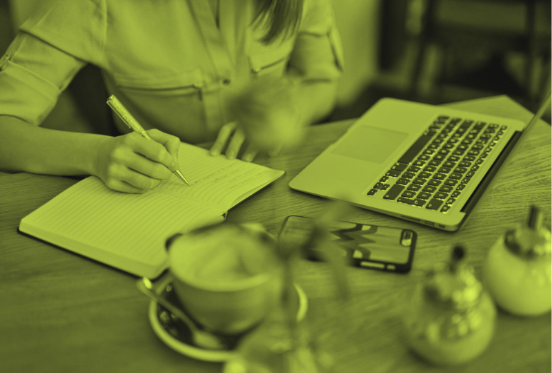 Person writing in a notebook at a wooden table with a laptop, smartphone, and condiments.